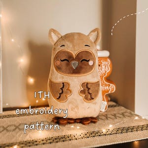 May include: A plush owl toy with light brown fur, dark brown facial markings, and embroidered closed eyes. The owl features small ears, two brown wings, and brown feet. The text ITH embroidery pattern is visible on the left side of the plush toy.