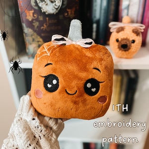 May include: A plush orange pumpkin toy with a grey stem, a light pink ribbon, and embroidered facial features including large black eyes, a smile, and pink cheeks. A spider web is stitched on the side. Text reads ITH embroidery pattern.