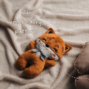 Op de afbeelding: Een pluche vos met oranje en witte vacht, voorzien van geborduurde ogen, een zwarte neus en een blauw-witte gingham strik. De tekst ITH embroidery pattern is erboven zichtbaar. Het speelgoed heeft een zachte, pluizige textuur.