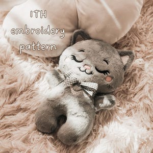 Könnte beinhalten: Ein graues Plüschkatzenspielzeug mit gestickten geschlossenen Augen, einer rosa Nase und einer kleinen karierten Fliege am Hals. Der Text ITH embroidery pattern ist über der Katze sichtbar. Das Plüschtier hat weiches Fell und gestickte Gesichtsdetails.