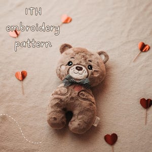 May include: A brown plush bear toy featuring embroidered facial details, a small black and white checkered bow tie at the neck, and a heart-shaped patch on the chest. The text "ITH embroidery pattern" is visible in the upper left corner.