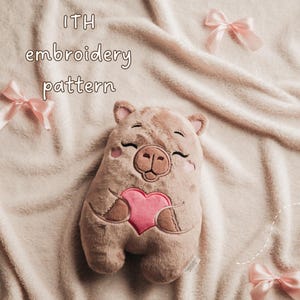 May include: A plush toy shaped like a pig with brown fur, embroidered facial features, and a pink heart on its chest. The text ITH embroidery pattern is visible. The toy is surrounded by light pink satin ribbons on a soft, textured beige fabric.