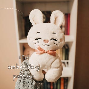 Op de afbeelding: Een witte pluche konijn met geborduurde details, waaronder een lachend gezicht en snorharen. Het heeft een roze strik met witte stippen en de tekst "ITH embroidery pattern". Het konijn is zacht.