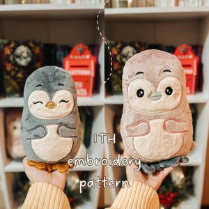 Può includere: Due peluche a forma di pinguino con il testo ITH embroidery pattern. Il pinguino a sinistra è grigio scuro con pancia color crema e piedi arancioni. Il pinguino a destra è marrone chiaro con pancia color crema e piedi grigi. Volti ricamati.