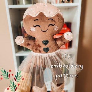 May include: A plush gingerbread person toy with brown fabric, a light pink icing-style head topping, and pink embroidered cheeks. It features two dark green heart-shaped buttons on the chest and a white tulle skirt. Text reads ITH embroidery pattern.