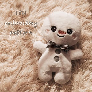 May include: A white plush snowman toy featuring embroidered black eyes with long lashes, a small orange nose, and pink cheeks. It wears a black and white chequered bow tie and has two grey circular buttons on its chest. The text ITH embroidery pattern is visible.