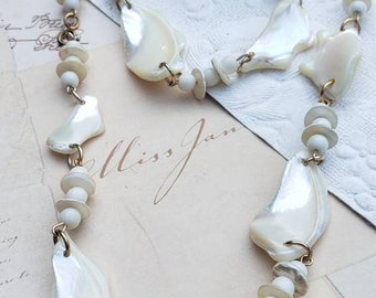 May include: A white shell necklace with gold accents and white beads. The necklace is on a white surface with a handwritten note that says "Miss Jan".