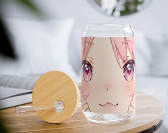 Op de afbeelding: Een helder glazen beker in de vorm van een blikje met een rozeharig anime-personagegezicht en grote paarse ogen. Het heeft een bamboe deksel en een doorzichtig rietje. De beker staat op een houten tafel.