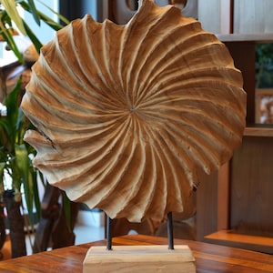 May include: A wooden, spiral-carved sculpture, mounted on a wooden base with metal supports. The sculpture is a light brown color and sits on a wooden table. The background includes other wooden decor.