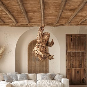 May include: A hanging wooden sculpture of a person, crafted from various pieces of wood, suspended from a ceiling beam. The sculpture is light brown and detailed, with arms raised. The room features a white sofa, arched doorway, and wooden door.