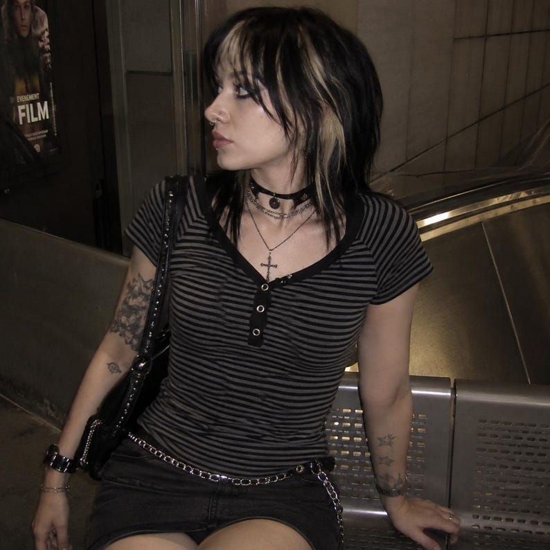 May include: A short-sleeved henley top featuring horizontal black and grey stripes, a black button placket, and a V-neckline. The top is worn with a black skirt, a black shoulder bag, a layered necklace with a cross pendant, and a choker.