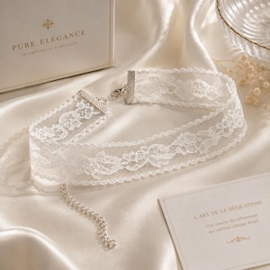 May include: A white lace choker necklace with a delicate floral pattern. The necklace has a silver clasp and a small chain. The words "PURE ELEGANCE" and "INTEMPORELLE & DELICATE" are printed on a box in the background.