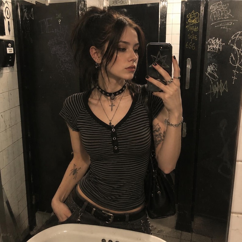 May include: A short-sleeved, V-neck henley top featuring thin black and dark grey horizontal stripes. The top has a buttoned placket down the centre chest. The garment is paired with a black belt and a black shoulder bag.