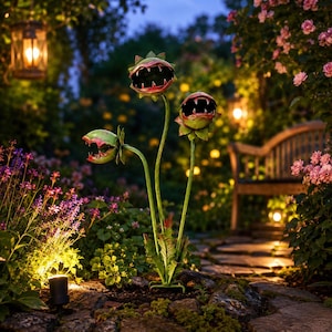 May include: A garden sculpture featuring three green, plant-like heads with open, toothy mouths and red accents. The heads are mounted on thin, upright stems rising from a base of green leaves, creating a tall, multi-headed botanical figure.