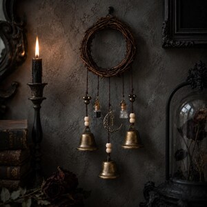 Puede incluir: Un adorno de pared que consiste en una corona circular de ramas con tres campanas de lat&oacute;n colgantes, dos peque&ntilde;os frascos de vidrio con contenido oscuro y claro, cuentas de madera y un dije de media luna de metal central.
