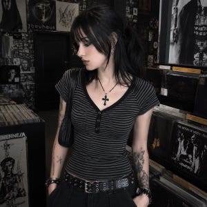 May include: A short-sleeved, V-neck henley shirt featuring horizontal black and dark grey stripes. The shirt has a button-down placket at the centre chest. The garment is paired with a black belt featuring two rows of metal grommets and a black shoulder bag.