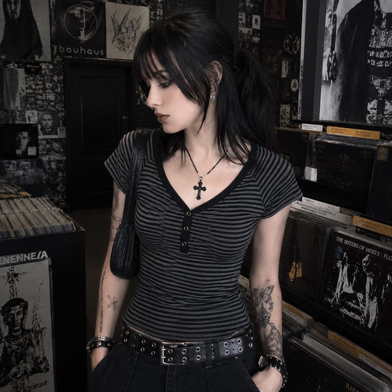 May include: A short-sleeved, V-neck henley shirt featuring horizontal black and dark grey stripes. The shirt has a button-down placket at the centre chest. The garment is paired with a black belt featuring two rows of metal grommets and a black shoulder bag.