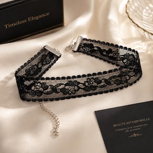 May include: Black lace choker necklace with floral patterns and a silver-tone clasp. The choker is displayed on a white satin surface. A black box with the words "Timeless Elegance" is in the background.