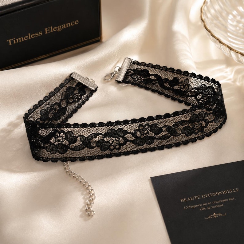 May include: Black lace choker necklace with floral patterns and a silver-tone clasp. The choker is displayed on a white satin surface. A black box with the words "Timeless Elegance" is in the background.