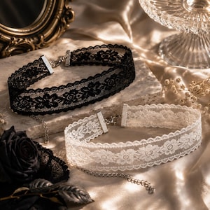 May include: Two lace chokers, one black and one white, with floral patterns. Each choker has a silver-toned clasp and chain. The chokers are displayed on a neutral surface with a satin-like background, suggesting a fashion or accessory theme.