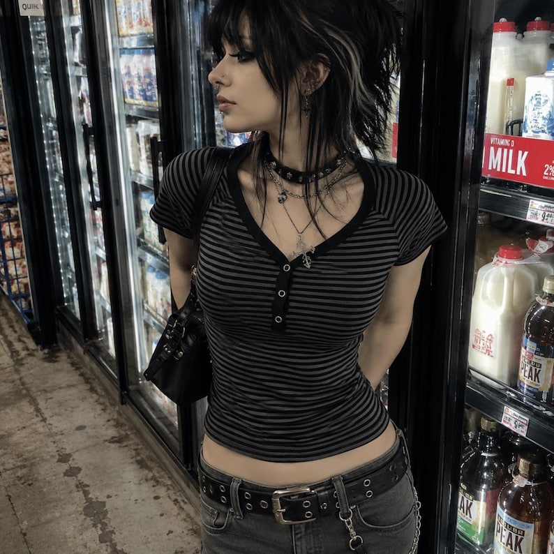 May include: A short-sleeved, V-neck henley shirt featuring thin horizontal black and grey stripes, a button-down placket, and a fitted silhouette. The shirt is paired with a black leather belt with silver-toned grommets and a black shoulder bag.