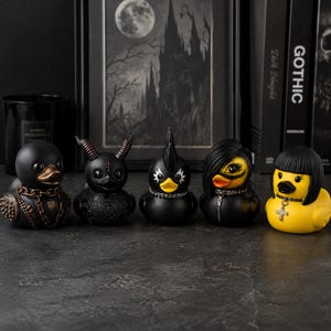 Puede incluir: Una colección de cinco patos de goma con diseños distintos: uno con cadena dorada y pinchos, uno con cuernos y un pentagrama, uno con cresta, uno con pelo largo negro y un pato amarillo con corte de pelo bob y un collar con una cruz.