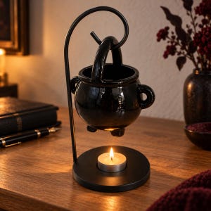 May include: A black ceramic cauldron-shaped wax warmer suspended from a curved metal stand. The glossy, round vessel features two side handles and a top handle, resting above a circular base that holds a small, lit white tea light candle.