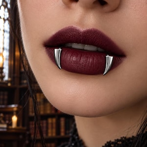 Könnte beinhalten: Ein Paar silberfarbene, fangzahnförmige Lippenpiercings an der Unterlippe. Der Schmuck hat eine glatte, polierte Oberfläche und ein geschwungenes, spitzes Design, das das Aussehen von Vampirzähnen vor einem dunklen Lippenstift nachahmt.