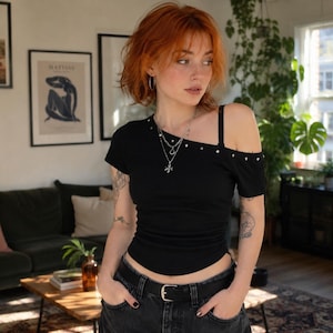 Puede incluir: Una camiseta negra de manga corta con escote asimétrico, un tirante y tachuelas plateadas a lo largo de la clavícula. La prenda se combina con unos vaqueros oscuros sujetos por un cinturón negro con una hebilla plateada.