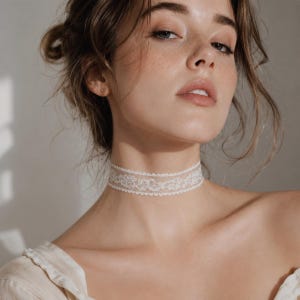 May include: A delicate, white lace choker necklace. The choker features an intricate floral pattern and is worn around the neck. The background is a soft, neutral color, highlighting the necklace's details.