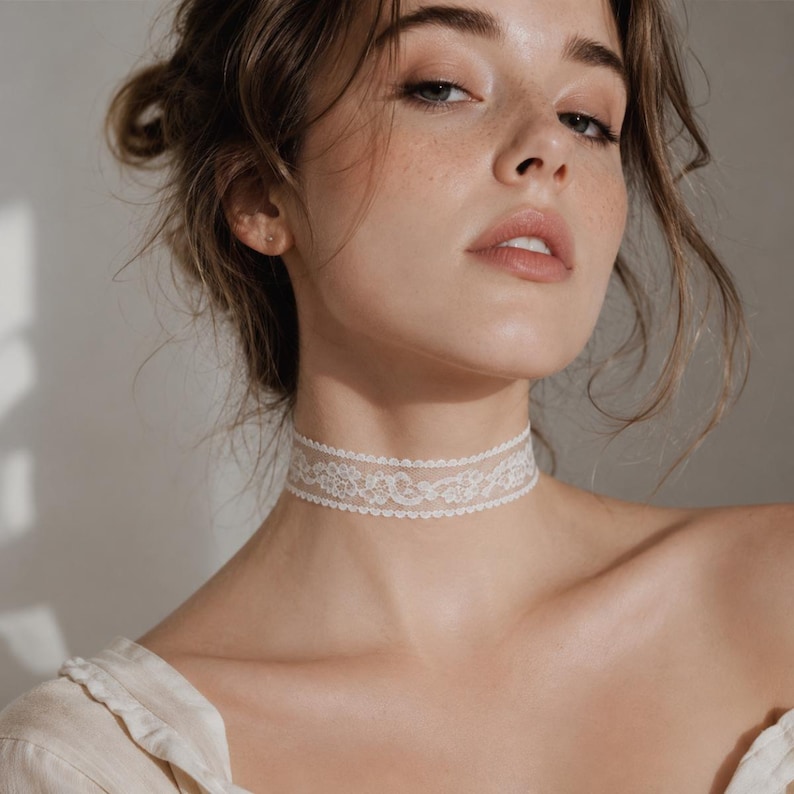 May include: A delicate, white lace choker necklace. The choker features an intricate floral pattern and is worn around the neck. The background is a soft, neutral color, highlighting the necklace's details.