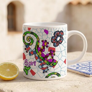 May include: White ceramic mug featuring a colorful lizard design with the word "Spirit". The lizard is composed of various colors, including green, purple, and yellow. The mug has a white handle and is decorated with a floral and geometric pattern.