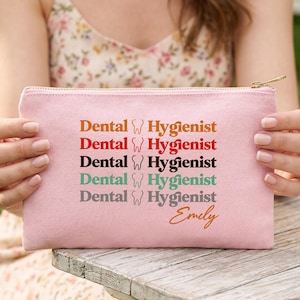May include: A light pink canvas pouch with a gold zipper. The text "Dental Hygienist" is printed in multiple colors. The name "Emily" is printed in a cursive font. The pouch is held in front of a wooden table.
