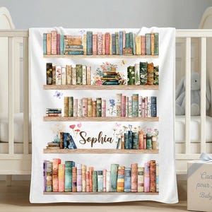 Puede incluir: Una manta de bebé blanca con un diseño de acuarela de estanterías llenas de libros coloridos. El nombre "Sophia" está escrito en cursiva. Mariposas y flores adornan las estanterías. La manta está sobre una cuna blanca.