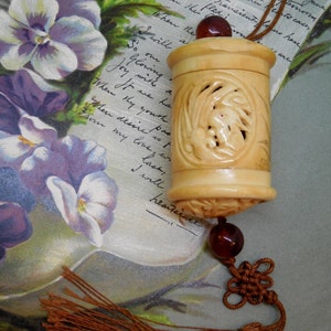 May include: A cylindrical, cream-colored carved pendant featuring intricate relief patterns, topped with a dark reddish-brown bead. A matching bead connects the pendant to a decorative brown cord knot and a long, flowing brown fabric tassel.