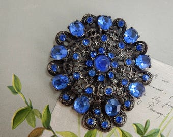 May include: A round brooch featuring a dark metal filigree base adorned with numerous blue faceted crystals of varying sizes. The design includes a large central stone surrounded by smaller stones arranged in a circular pattern.
