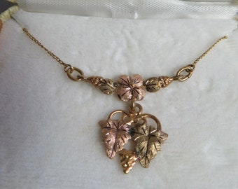 May include: A tri-color gold necklace featuring a central flower motif and a dangling grape leaf pendant. The jewelry is presented in a cream-colored box with the gold-printed text Black Hills Creations by Lundstrom on the interior lid.