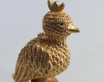 May include: A gold-toned metal bird figurine featuring a textured feather pattern, a small crest on its head, and a single dark eye. The bird is mounted on a circular, tiered pedestal base with a matching textured finish and a pointed beak.