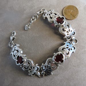 Pu&ograve; includere: Un bracciale a maglie in argento con segmenti metallici decorati da volute che si alternano a pietre rosso scuro a forma di cuore. Il bracciale include una chiusura a moschettone e una catenella regolabile con un ciondolo a cuore.
