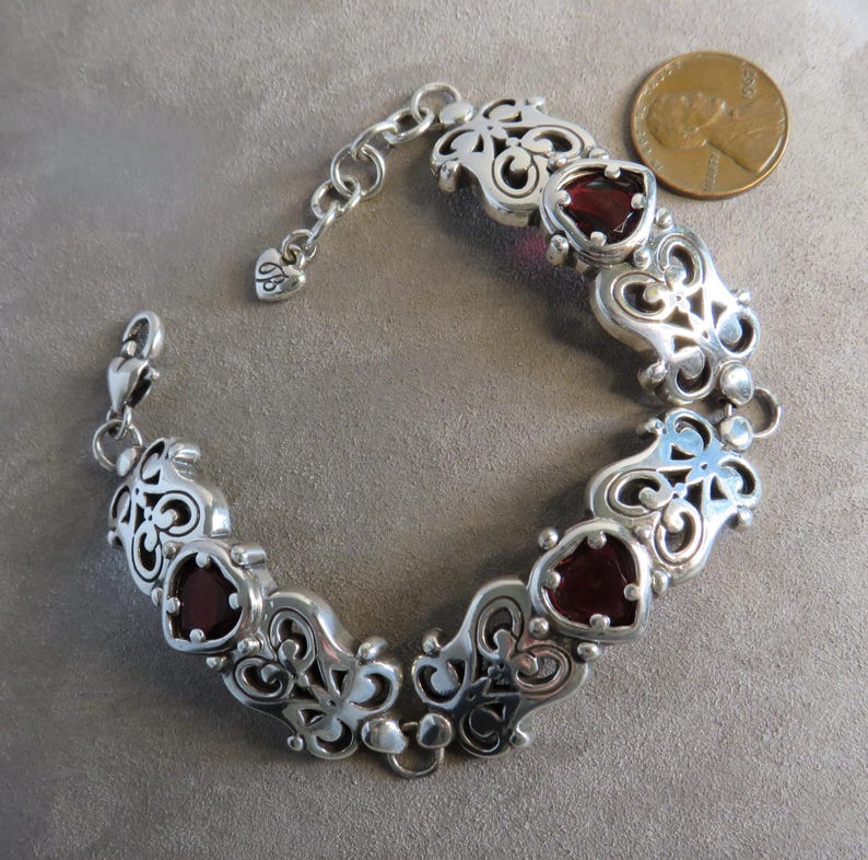 Pu&ograve; includere: Un bracciale a maglie in argento con segmenti metallici decorati da volute che si alternano a pietre rosso scuro a forma di cuore. Il bracciale include una chiusura a moschettone e una catenella regolabile con un ciondolo a cuore.