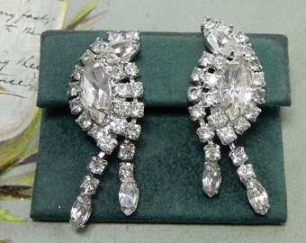 May include: A pair of silver-toned earrings featuring clear marquise and round-cut rhinestones arranged in a curved, leaf-like cluster design with two dangling, faceted stone drops extending from the base of each earring.