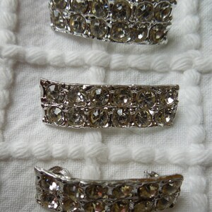 May include: Three rectangular silver-toned metal accents, each featuring two horizontal rows of clear, faceted circular rhinestones. The pieces have a slightly curved, elongated shape and a metallic finish, arranged vertically in a column.