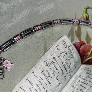 Peut inclure: Bracelet en argent avec des d&eacute;tails roses et des breloques, dont un c&oelig;ur et un ruban rose. Le bracelet est pr&eacute;sent&eacute; avec un livre ouvert et des fleurs, sugg&eacute;rant un th&egrave;me de soutien ou de souvenir.
