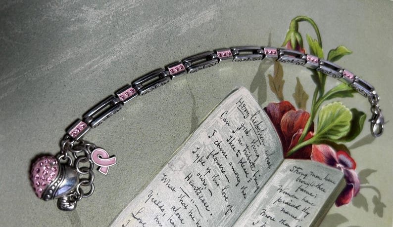 Peut inclure: Bracelet en argent avec des d&eacute;tails roses et des breloques, dont un c&oelig;ur et un ruban rose. Le bracelet est pr&eacute;sent&eacute; avec un livre ouvert et des fleurs, sugg&eacute;rant un th&egrave;me de soutien ou de souvenir.
