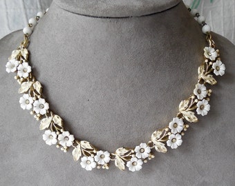 May include: A gold-tone metal necklace featuring a series of white enamelled flowers and leaves. The floral design is accented with small clear rhinestones at the centre of each blossom. The necklace is finished with a strand of white beads.