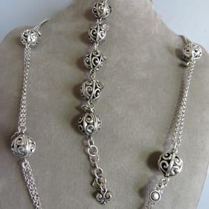 May include: A silver necklace and bracelet set with decorative, openwork, round beads. The necklace is longer than the bracelet and has a chain link design.