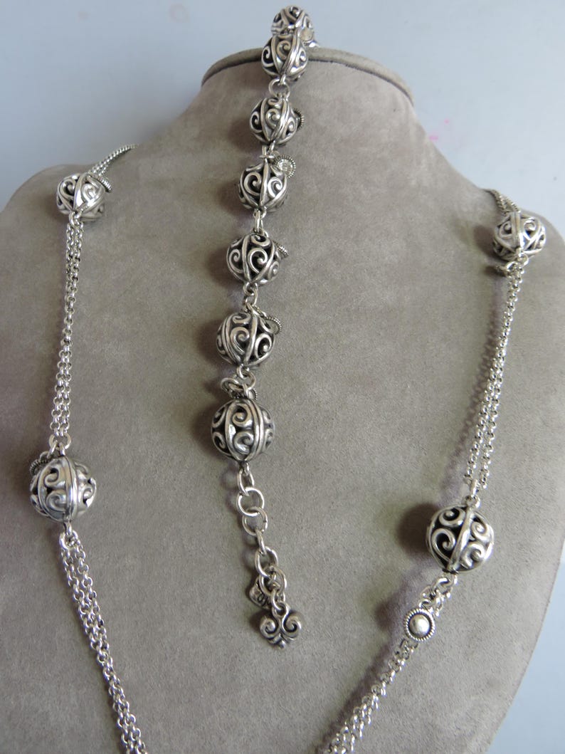 May include: A silver necklace and bracelet set with decorative, openwork, round beads. The necklace is longer than the bracelet and has a chain link design.