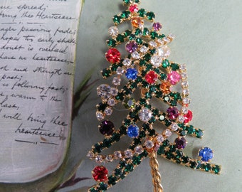 May include: A gold-tone Christmas tree brooch with green, red, blue, and clear rhinestones. The tree is decorated with a variety of colorful gems and has a twisted gold wire base.