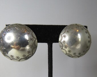 May include: A pair of circular silver earrings with a polished, domed surface. The edges of each earring are decorated with a stamped pattern consisting of small star-like shapes and a continuous zigzag line encircling the perimeter.