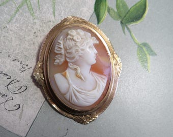 May include: Oval cameo brooch with a profile of a woman in white relief against an orange background. The cameo is set in a gold-colored frame with floral detailing. The brooch is a vintage piece.